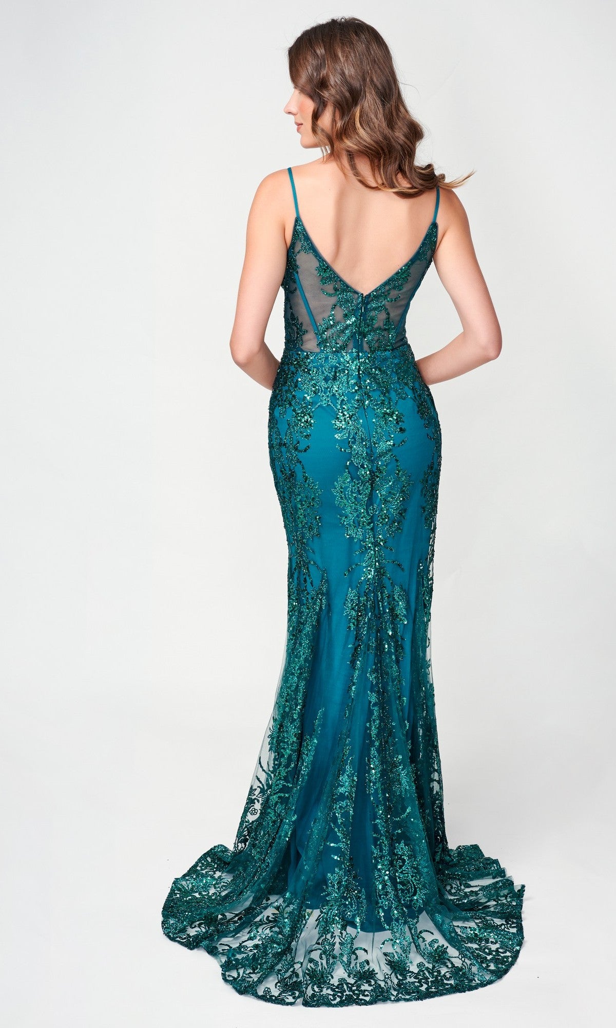 A woman stands facing away in front of a plain light background, wearing the Nina Canacci 1562—a teal, sleeveless, floor-length mermaid dress featuring lace, sequin detailing, and hot stone embellishment.